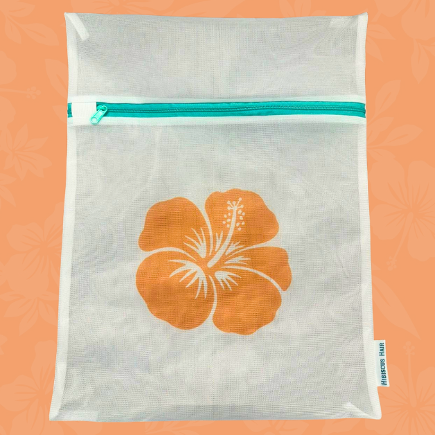 A white mesh laundry bag with a teal zipper and a large orange hibiscus flower printed on the front, set against a peach background with a subtle floral pattern.