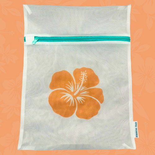 A white mesh laundry bag with a teal zipper and a large orange hibiscus flower printed on the front, set against a peach background with a subtle floral pattern.