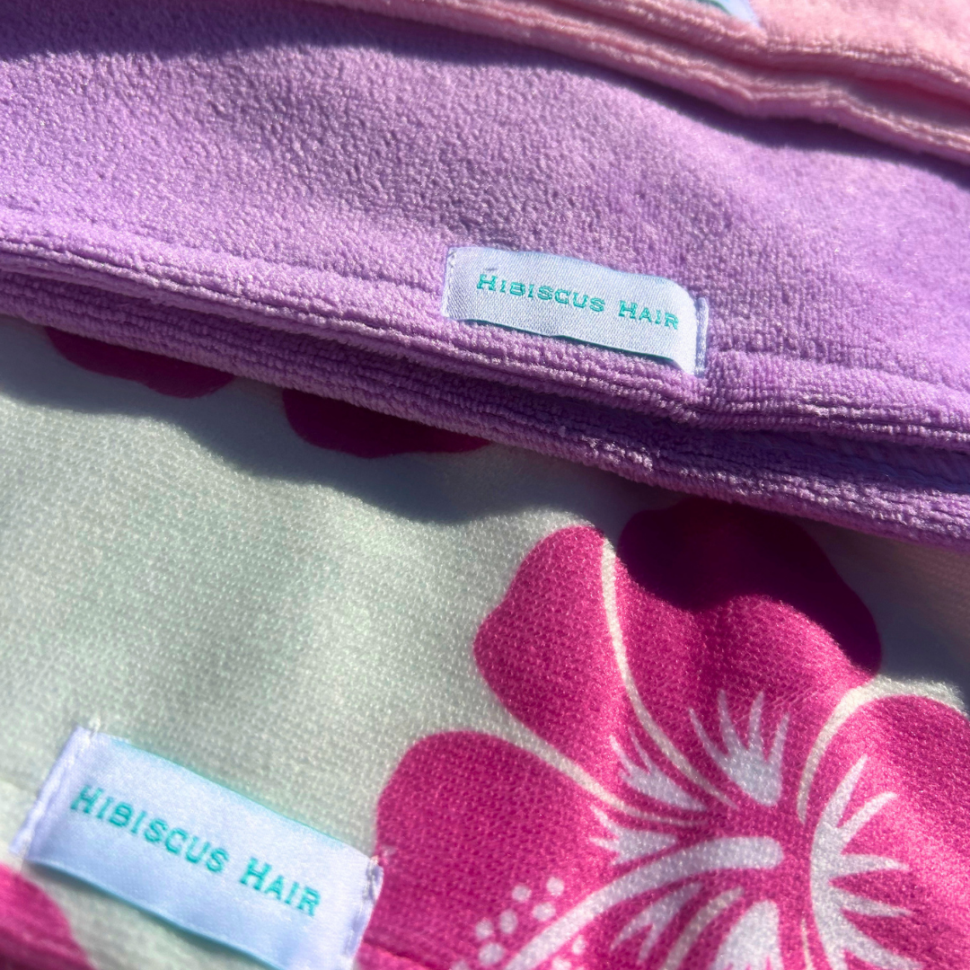 Close-up of microfibre hair wraps with visible 'HIBISCUS HAIR' labels. The top piece is solid lavender-purple, and the lower piece has a cream background with a large hot pink hibiscus flower print.