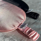 A close-up of a satin sleep mask in light pink with a matching ruched elastic strap, with a black sleep mask partially visible behind it on a textured gray surface.