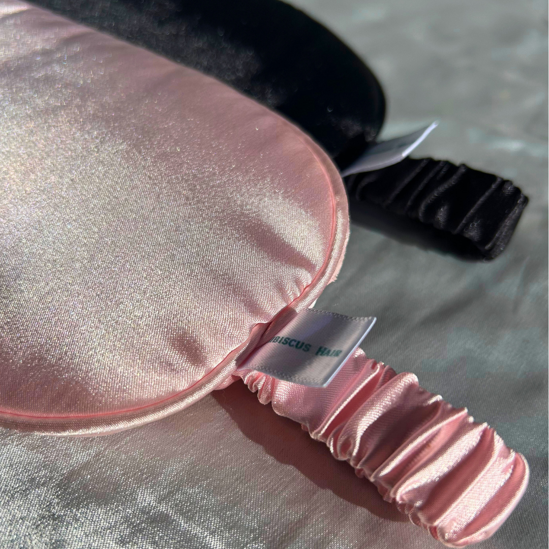 A close-up of a satin sleep mask in light pink with a matching ruched elastic strap, with a black sleep mask partially visible behind it on a textured gray surface.
